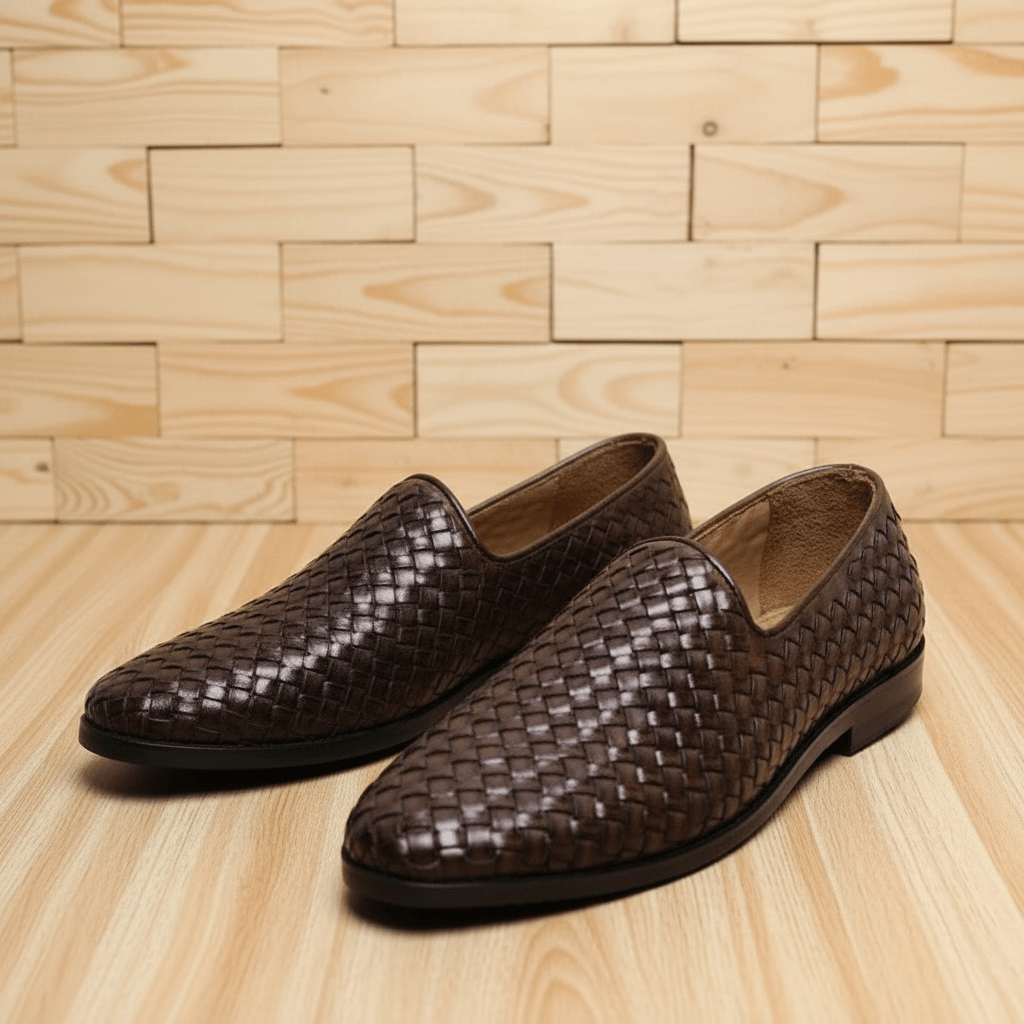 Pair of men's woven leather loafers in dark brown, displayed on a wooden surface, showcasing craftsmanship and classic design.